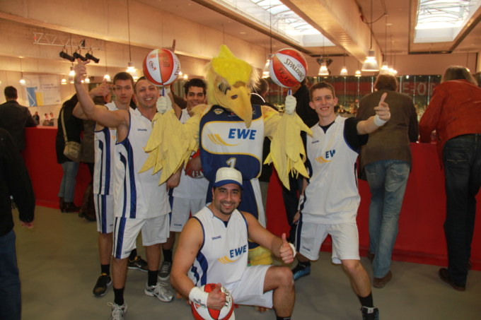 Dieses Bild zeigt fünf engagierte Breakballteam-Mitglieder, die um ein dynamisches Maskottchen gruppiert sind, welches zwei Bälle in einer faszinierenden Drehbewegung präsentiert – ein magisches Highlight der EWE Playoffs 2013, inszeniert von YOPURACT & Breakball®.